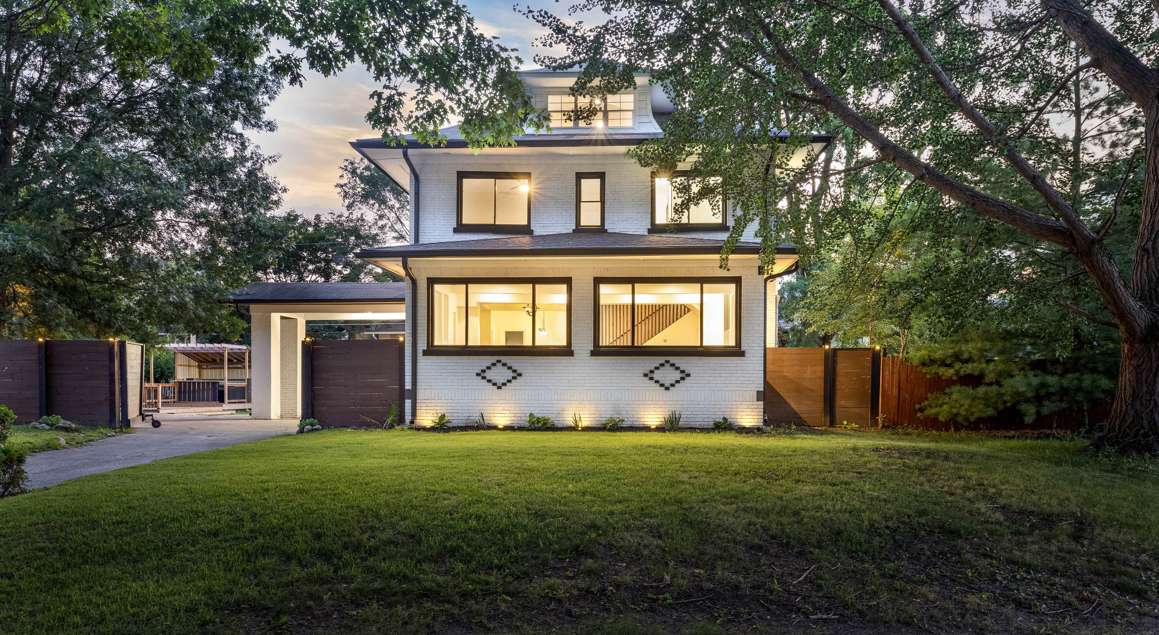 Illuminated white residential exterior framed by mature trees at twilight